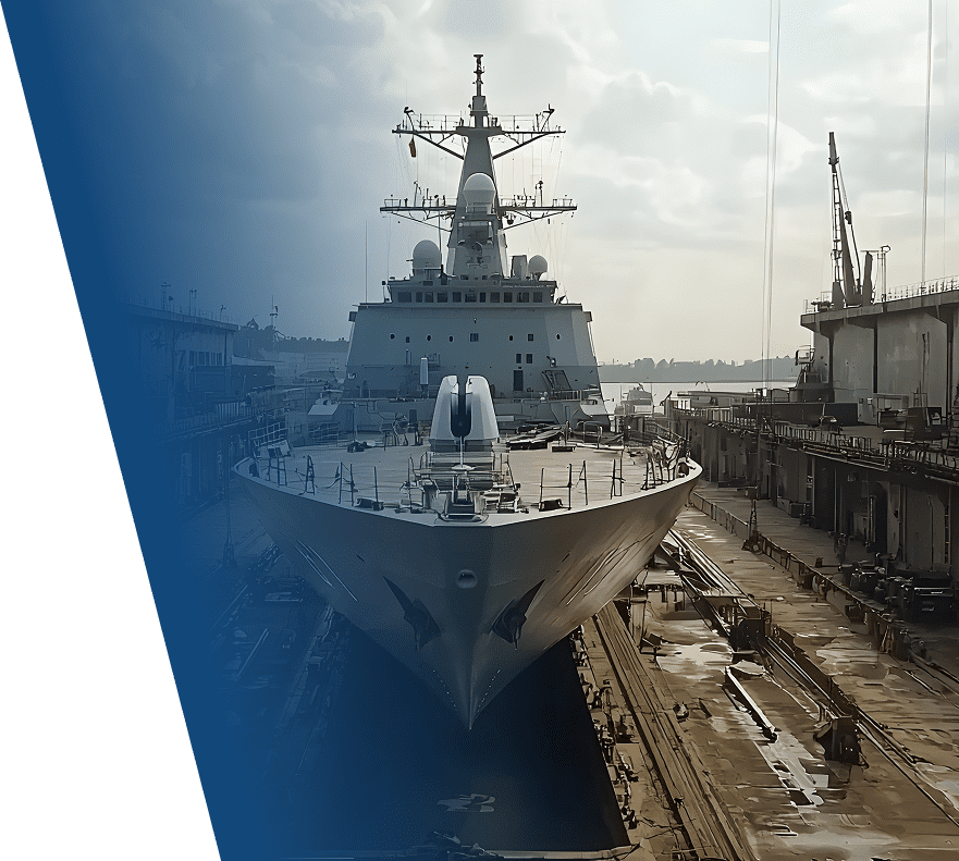 A large naval ship docked in a dry dock, viewed from the front. The ship has a prominent bow, multiple antennas, radar domes, and communication equipment on its superstructure. The dock area around the ship is industrial, with cranes and maintenance platforms visible. The lighting suggests early morning or late afternoon with a partly cloudy sky.