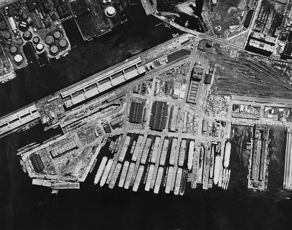 Aerial black-and-white photograph of a large industrial waterfront area featuring multiple docks with numerous ships and boats moored. The site includes several warehouses, storage tanks, and rail tracks, indicating a busy port or shipyard. The water surrounds the docks, and various buildings and infrastructure are visible throughout the area.