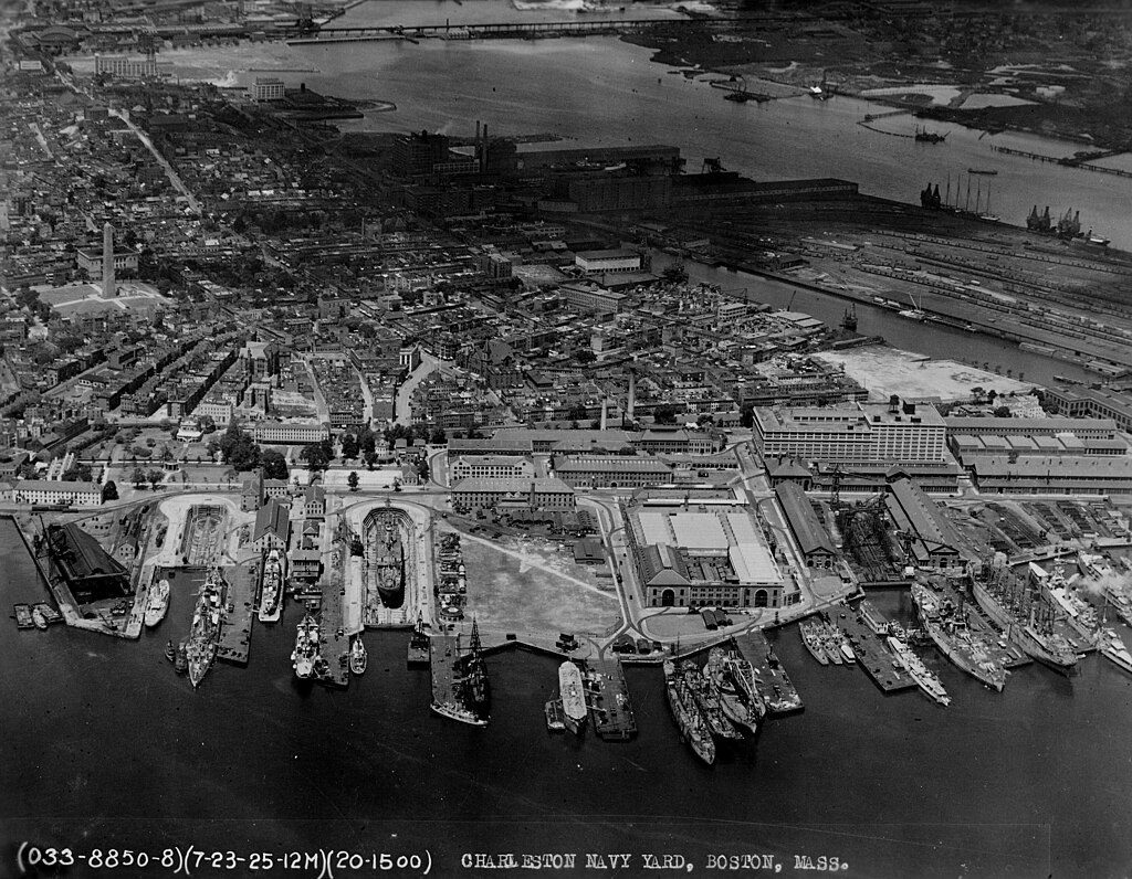 Aerial view of the Charleston Navy Yard in Boston, Massachusetts, showing multiple docks with naval ships moored, large industrial buildings, warehouses, and a dense urban area extending inland. The waterfront is busy with ships and cranes, and the image is in black and white, indicating a historical photograph.