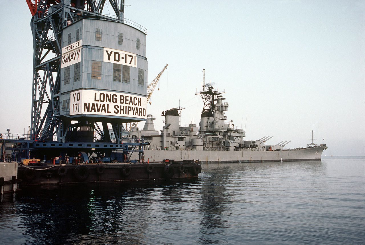 A large gray naval warship docked at a pier with a massive crane structure labeled "LONG BEACH NAVAL SHIPYARD," "YD-171," and "TITAN II GO NAVY." The ship has multiple gun turrets and radar equipment, and the water is calm with a clear sky in the background.