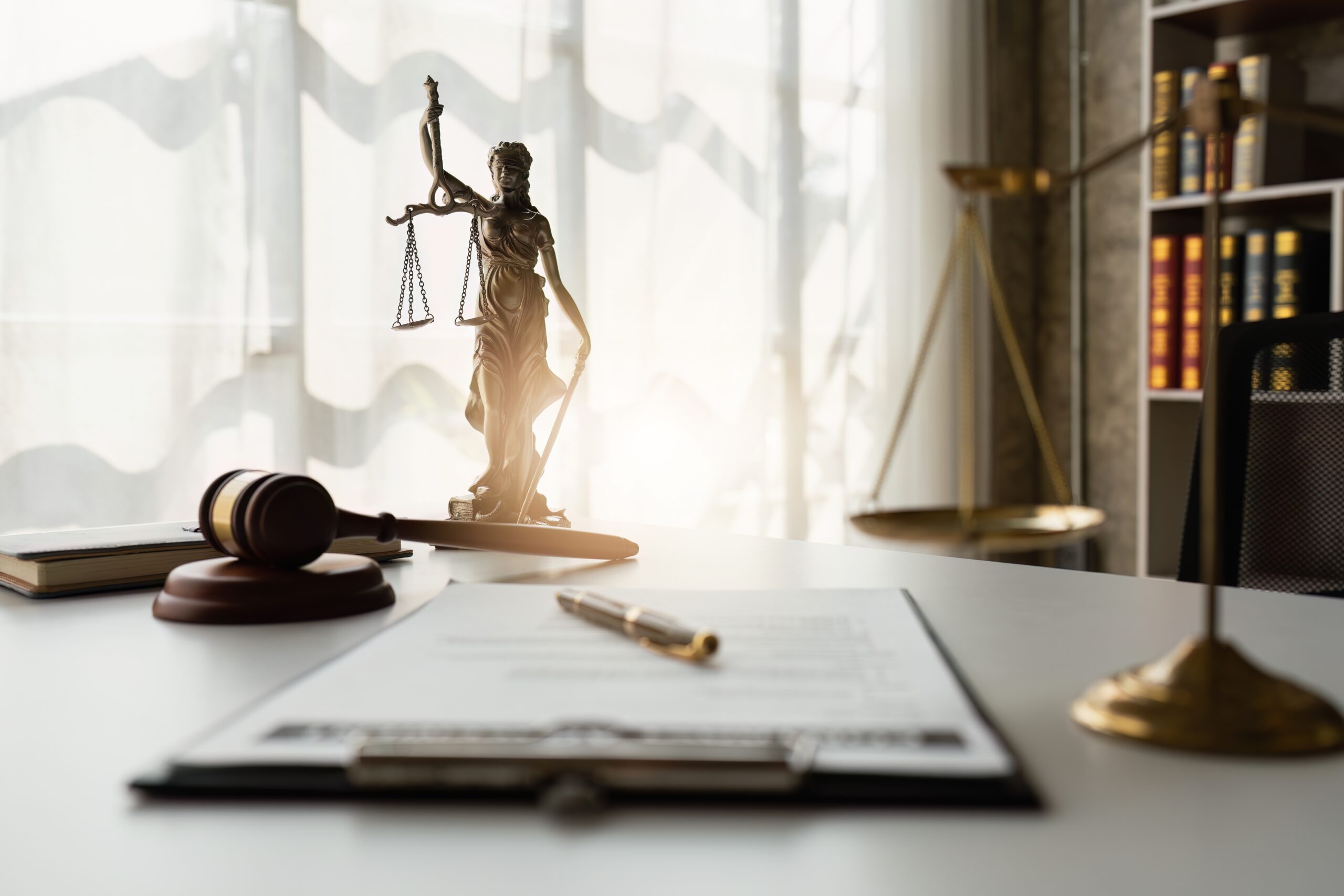 Bronze statue of Lady Justice holding scales and a sword, a wooden gavel on a sound block, a pen on a clipboard with documents, and a set of golden scales on a white desk in a law office with bookshelves in the background.