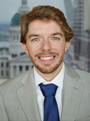 A young man with light brown hair and a beard is smiling. He is wearing a light gray suit jacket, a white dress shirt, and a textured dark blue tie. The background shows an urban cityscape with blurred buildings.
