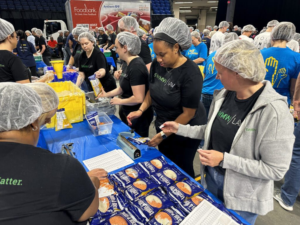 A group of volunteers wearing hairnets and black "SWMW | LAW" t-shirts are packing food items, including bags of rice and lentils, at a table covered with a blue plastic tablecloth. The table has sealing machines and printed sheets. In the background, more volunteers in blue shirts with yellow hand graphics are working, and a truck with "Foodbank St. Louis Area Foodbank" branding is visible.