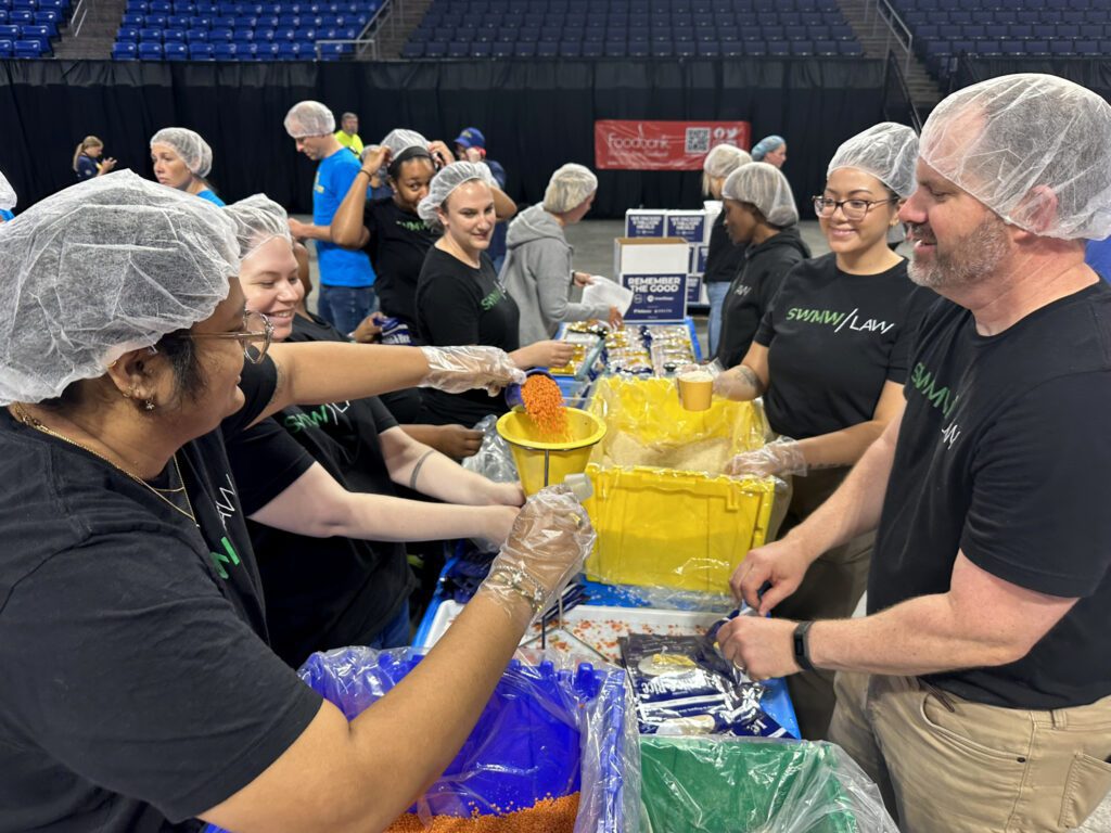 A group of volunteers wearing hairnets and gloves are working together at a packing station. They are scooping orange lentils or similar food into funnels to fill bags, which are then sealed. Most volunteers are wearing black shirts with "SWANN LAW" printed on them. The setting appears to be a large indoor venue with blue stadium seating in the background.