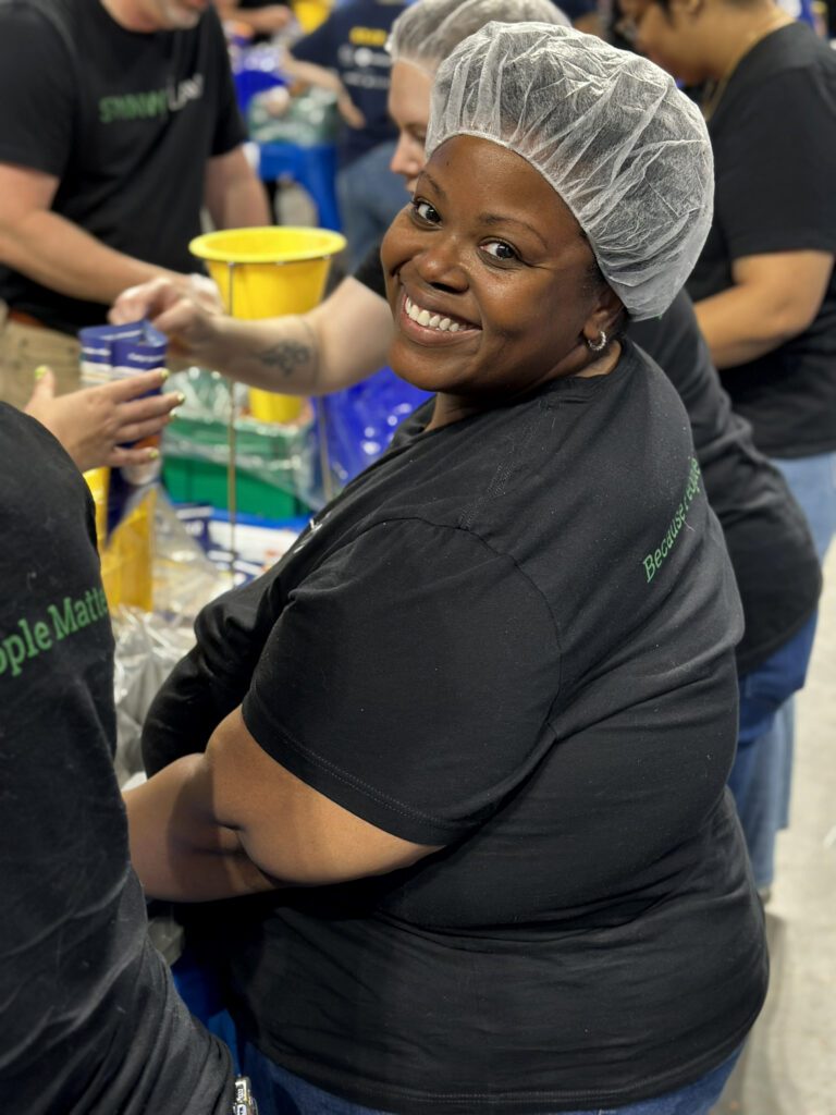 A smiling woman wearing a black t-shirt and a white hairnet is seated and looking over her shoulder at the camera. She is surrounded by other people who are also wearing black shirts and appear to be engaged in a group activity, possibly packaging or assembling items. The setting looks like an indoor workspace or volunteer event.