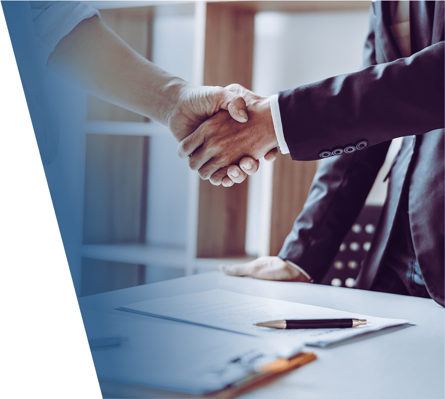 Two people shaking hands over a desk with documents and a pen, one person wearing a dark suit and the other in a light-colored shirt.