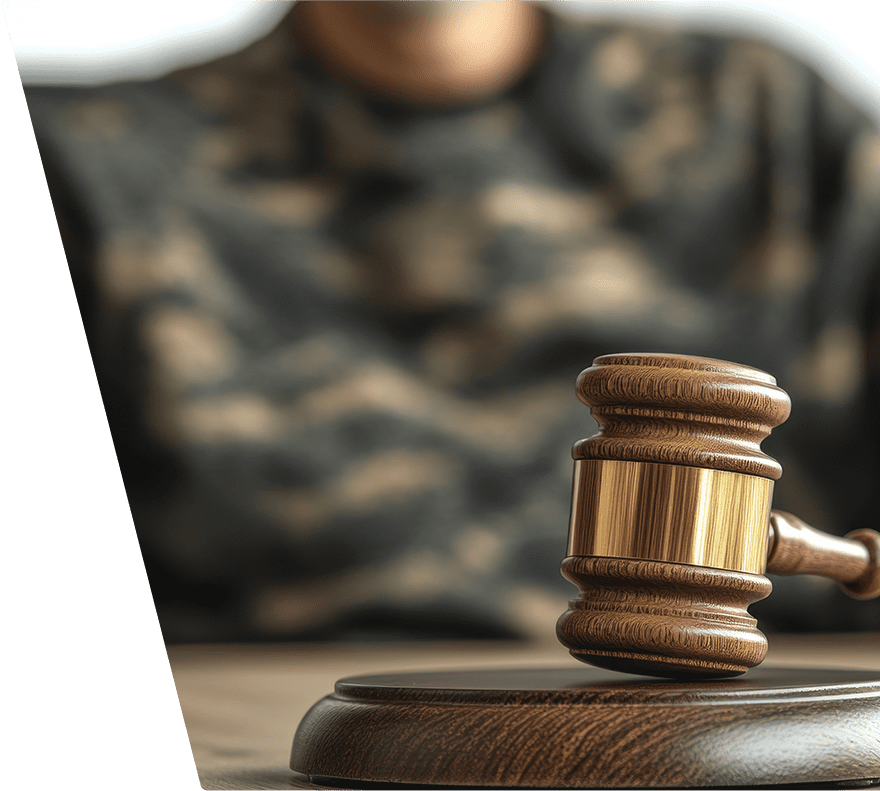 A wooden judge's gavel with a gold band rests on a matching wooden sound block, with a person wearing a camouflage-patterned shirt blurred in the background.