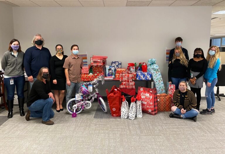 A group of eleven people wearing face masks are posing indoors around a table filled with wrapped Christmas gifts and a purple children's bicycle decorated with tinsel. The gifts are wrapped in various festive papers and gift bags, and the setting appears to be an office or similar environment with a neutral wall and carpeted floor.