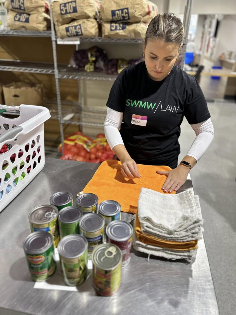 A woman wearing a black "SWMW / LAW" t-shirt and a name tag labeled "Emma" is folding orange and white towels on a metal table. In front of her on the table are several canned goods. In the background, there are metal shelves stocked with bags of produce and a white laundry basket filled with various items.