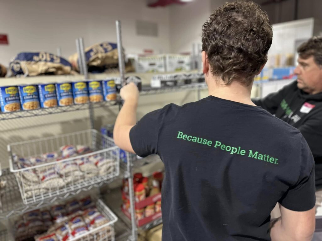 A person wearing a black t-shirt with the green text "Because People Matter." on the back is organizing canned goods on metal shelves. Another person is visible in the background, also engaged in organizing items. The shelves hold various food items, including canned beans and packaged goods.