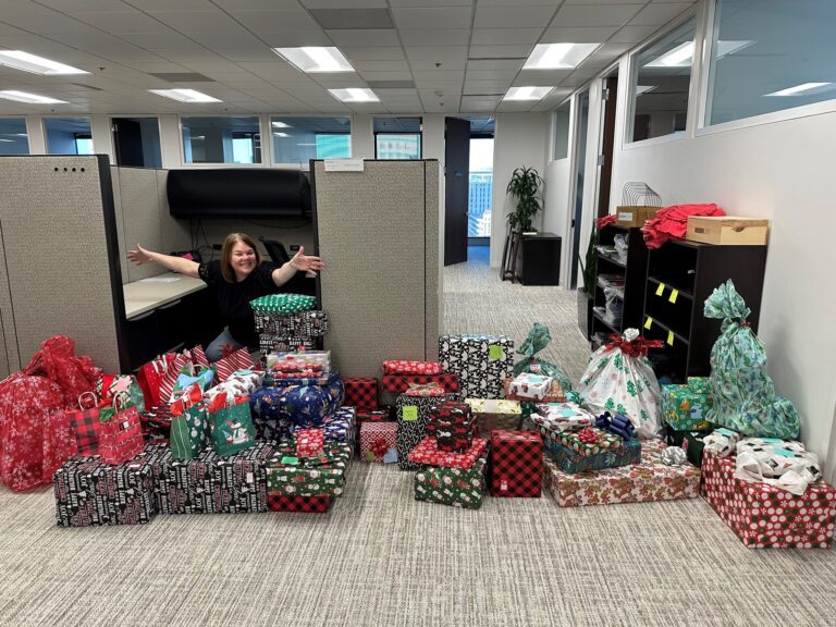 A woman with brown hair wearing a black shirt is smiling and extending her arms from inside an office cubicle. In front of her, there is a large collection of wrapped Christmas presents in various festive wrapping papers, including red, green, black, and white patterns with holiday motifs like snowflakes, snowmen, and Christmas trees. The office space has beige carpet, cubicle partitions, and shelves with folded red shirts and wooden boxes on the right side.