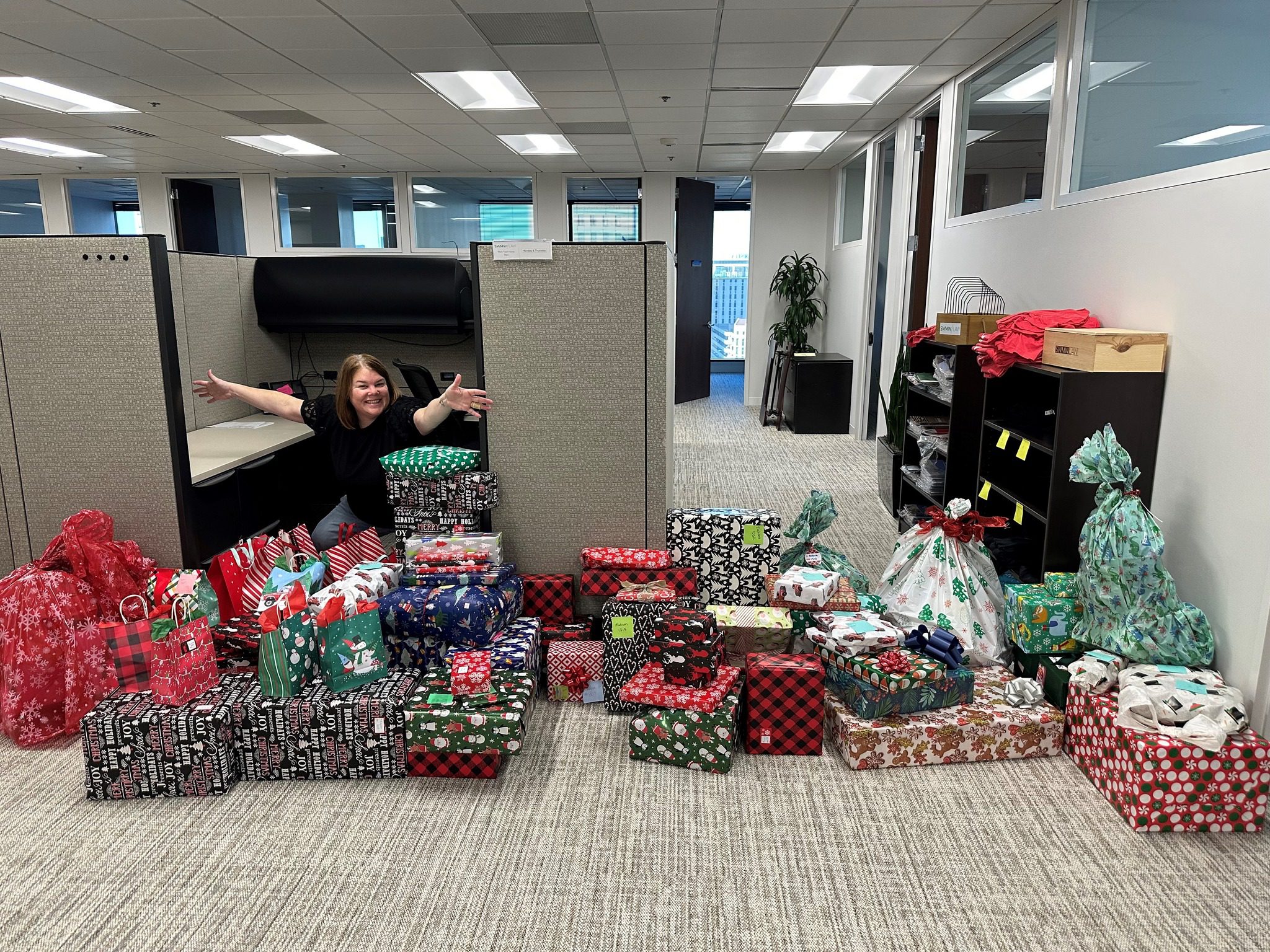 A woman with brown hair wearing a black shirt is smiling and extending her arms from inside an office cubicle. In front of her, there is a large collection of wrapped Christmas presents in various festive wrapping papers, including red, green, black, and white patterns with holiday motifs like snowflakes, snowmen, and Christmas trees. The office space has beige carpet, cubicle partitions, and shelves with folded red shirts and wooden boxes on the right side.