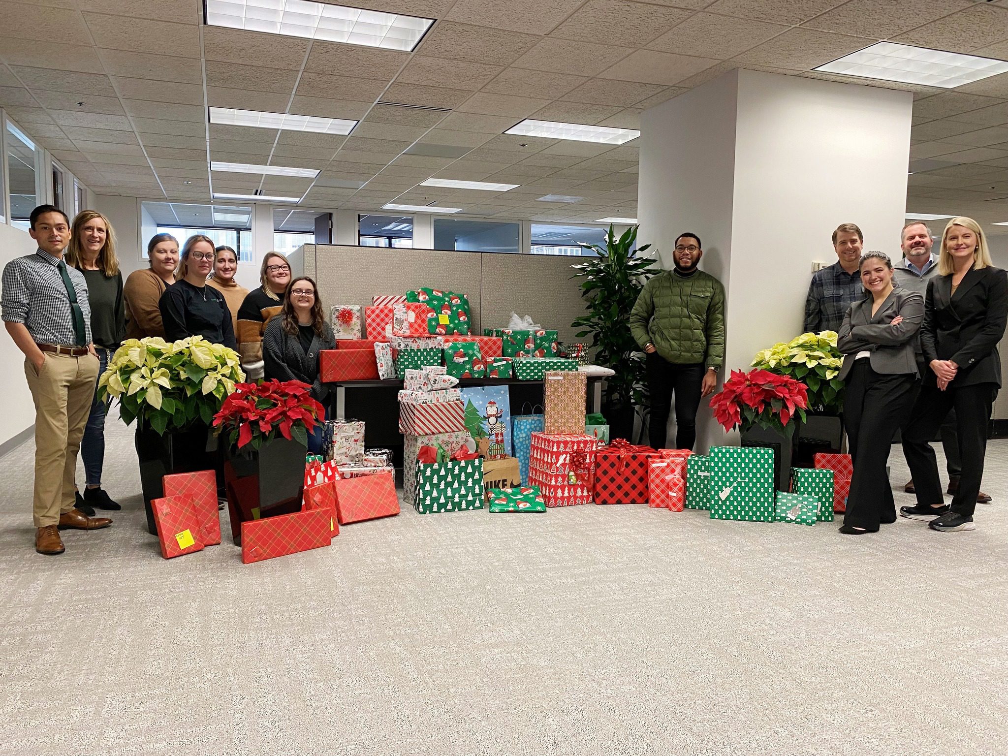 A group of twelve people standing in an office space behind a large collection of wrapped Christmas presents in various festive patterns and colors, including red, green, and white. There are also several pots of red and white poinsettias placed among the gifts. The office has a neutral color scheme with beige carpet and white walls.