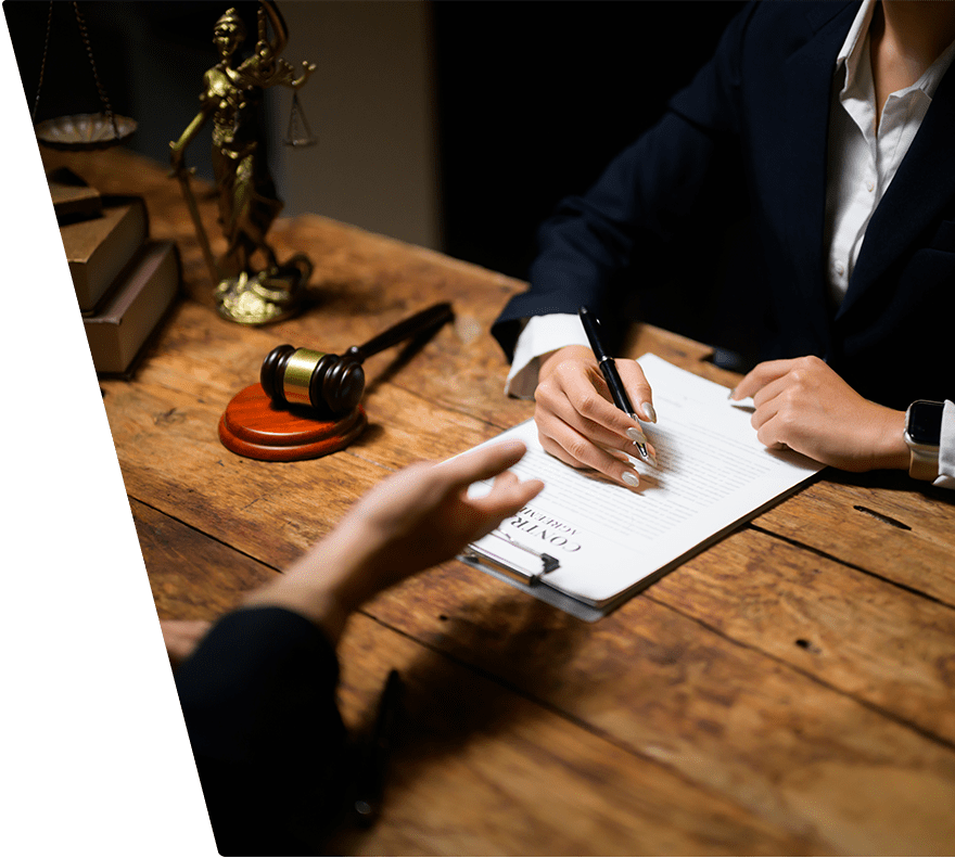 A person in a dark suit is holding a pen and reviewing a contract document on a clipboard, sitting at a wooden table. Across the table, another person is gesturing with their hand. On the table, there is a wooden gavel, a small statue of Lady Justice, and a stack of books.