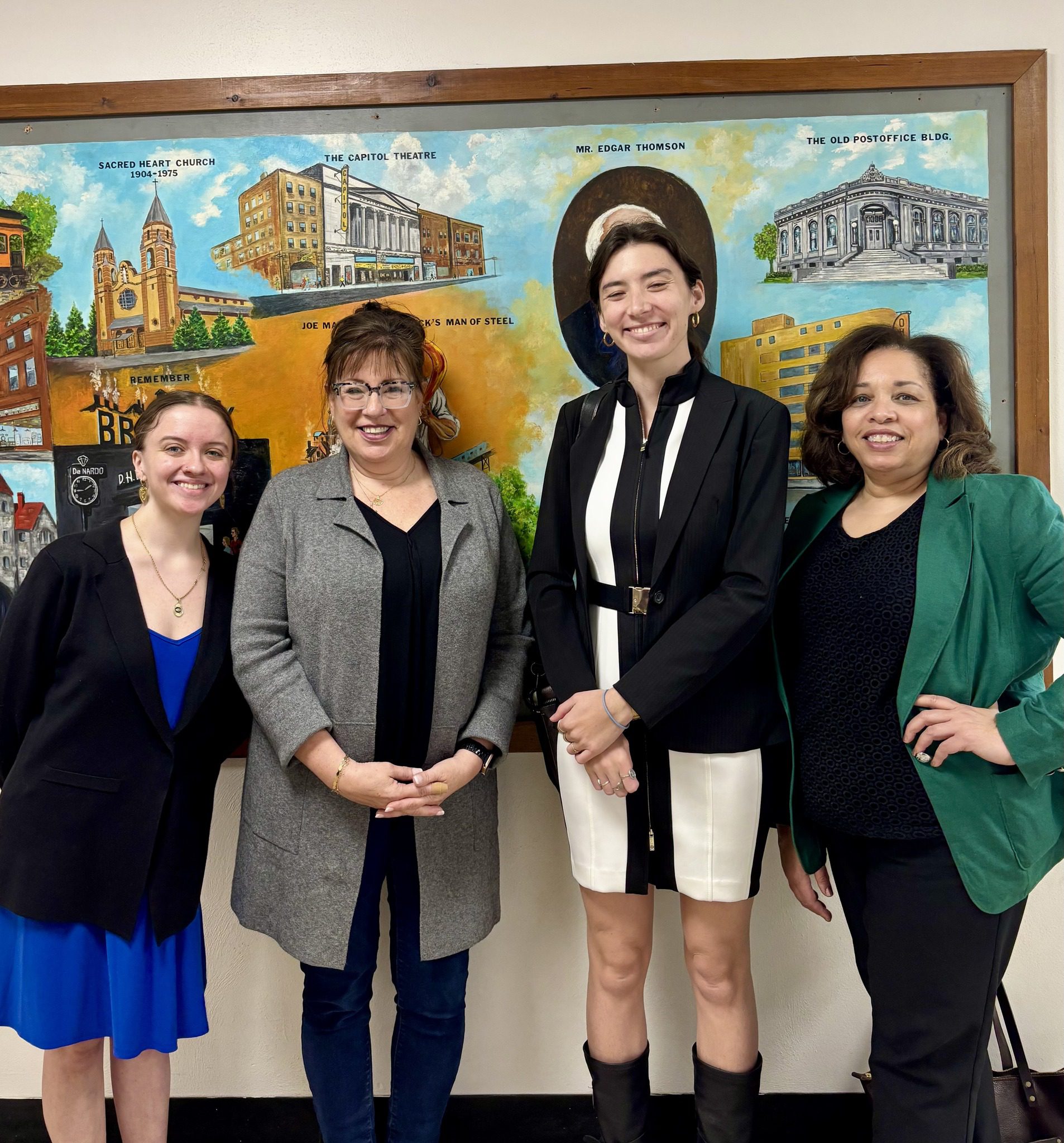 Four women stand smiling in front of a colorful mural depicting various buildings and landmarks, including Sacred Heart Church, The Capitol Theatre, and The Old Postoffice Building. The woman on the far left wears a blue dress with a black blazer, the second woman wears glasses, a gray coat, and dark pants, the third woman wears a black and white dress with a black blazer and black boots, and the woman on the far right wears a green blazer over a black top and black pants.