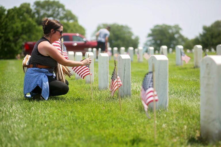 A woman wearing sunglasses, a black and white striped sleeveless top, black pants, and a blue jacket tied around her waist is kneeling on grass in a cemetery. She is placing small American flags next to white gravestones. In the background, another person is also placing flags among the gravestones. A red vehicle and trees are visible in the distance.