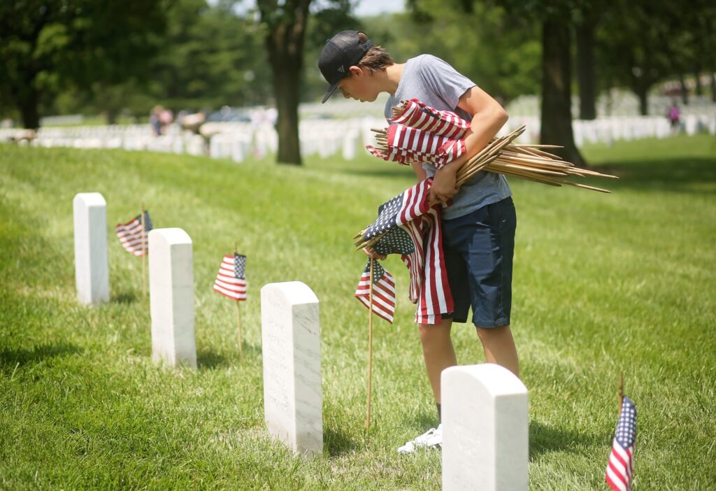 A young person wearing a gray t-shirt, navy shorts, white sneakers, and a black cap is placing small American flags on white gravestones in a cemetery. The individual is holding several flags in their arms while bending down to insert one into the ground. The scene is set on a sunny day with green grass and trees in the background.