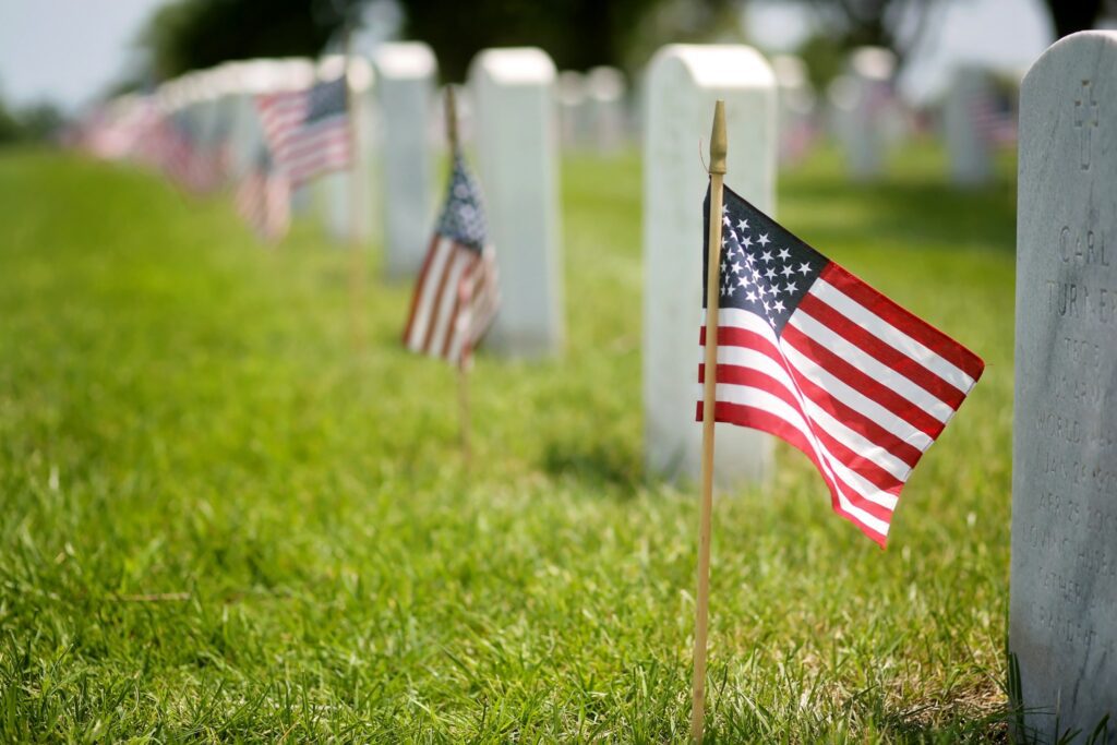 A row of white gravestones in a cemetery with small American flags placed in front of each one, set on green grass. The focus is on one flag and gravestone in the foreground, with the others fading into the background.