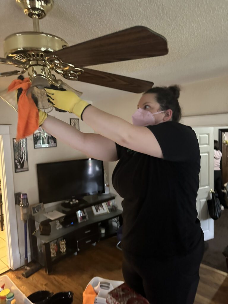 A person wearing a pink face mask and yellow gloves is cleaning a ceiling fan light fixture with an orange cloth in a living room. The ceiling fan has brass and wood elements. In the background, there is a TV on a stand with framed photos and decorative items. The room has wooden flooring and a vacuum cleaner is visible near the doorway.