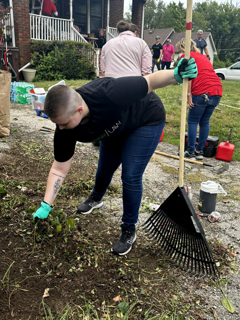 A person wearing a black t-shirt, blue jeans, and green gloves is pulling weeds or plants from the ground while holding a large black rake with a wooden handle. They have a short haircut and a tattoo on their left forearm. In the background, several other people are engaged in yard work near a brick house with a white porch railing. Various gardening tools, water bottles, and supplies are scattered around the area.