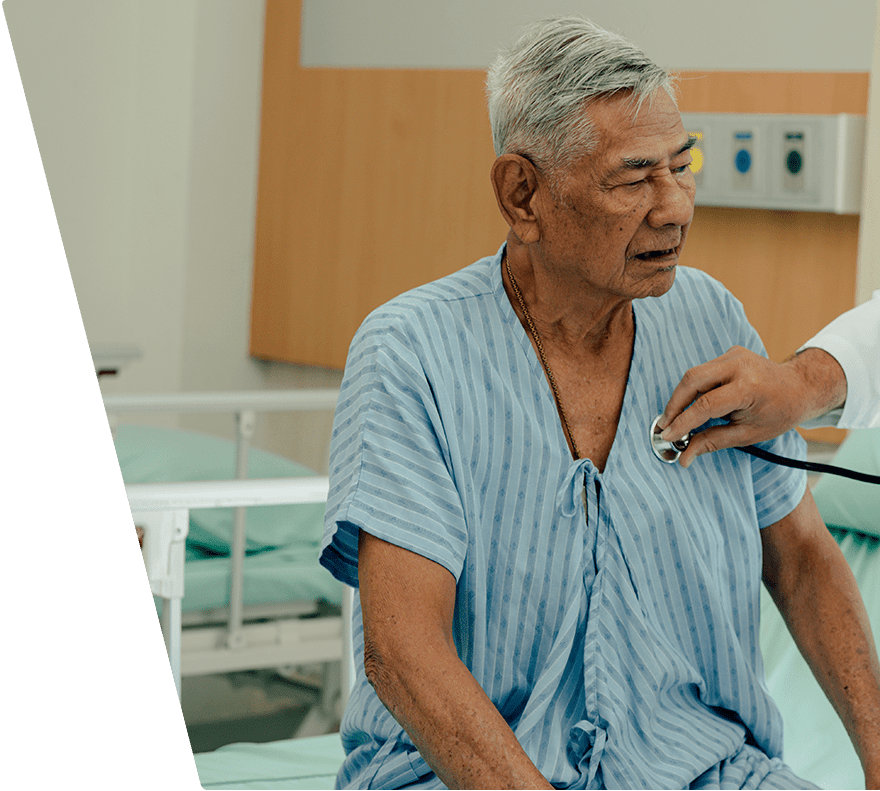An elderly man wearing a light blue striped hospital gown is sitting on a hospital bed while a healthcare professional uses a stethoscope to listen to his chest. The setting appears to be a hospital room with medical equipment and a bed in the background.