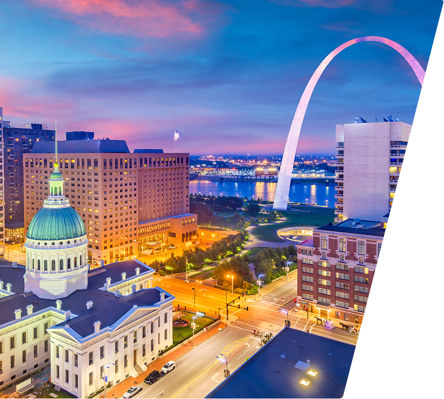 A cityscape at dusk featuring the illuminated Old Courthouse with a green dome in the foreground, modern buildings, and the iconic Gateway Arch in the background near a river. The sky is a gradient of blue and pink hues, and streetlights and building lights are glowing warmly.