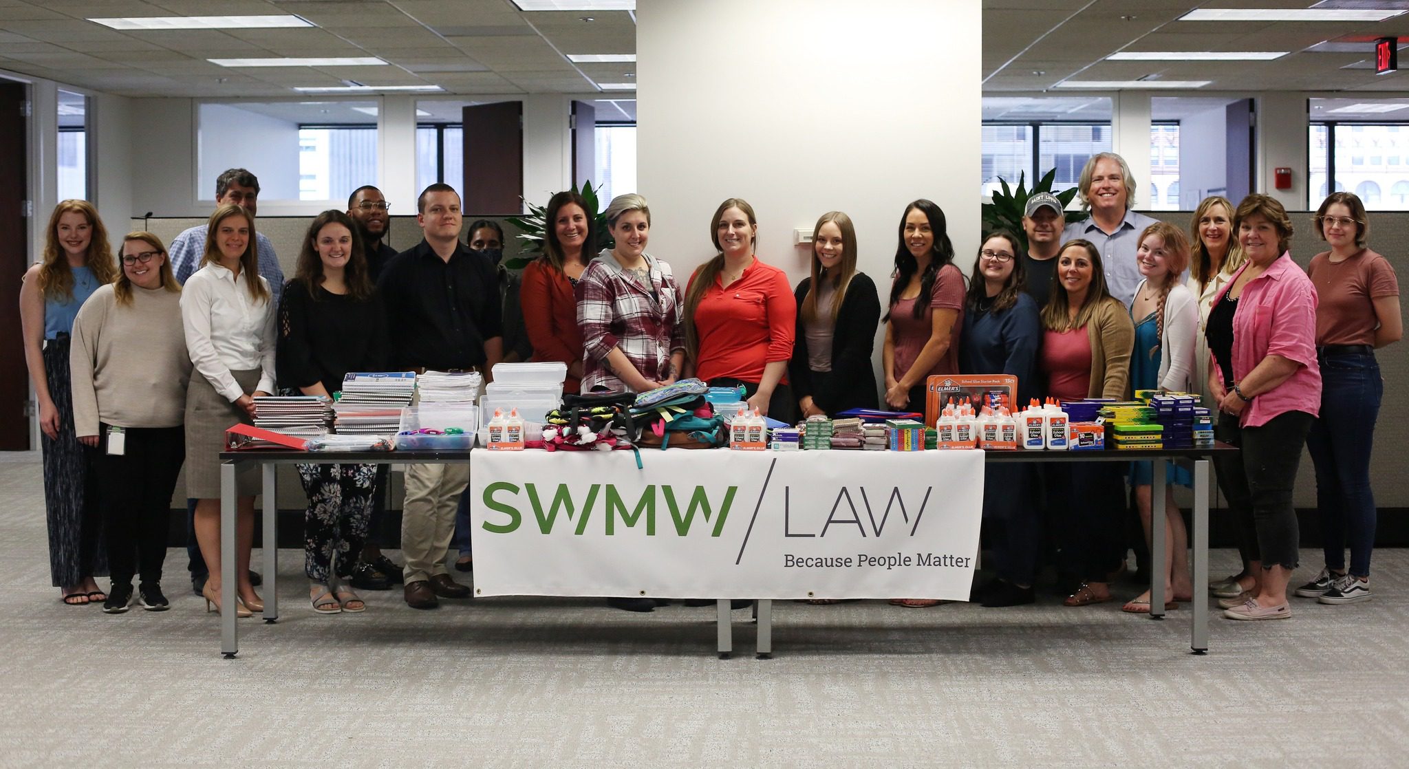 A group of people standing behind a long table filled with school supplies such as notebooks, glue bottles, pencils, and backpacks. A banner on the front of the table reads "SWMW LAW Because People Matter." The setting appears to be an office with cubicles and large windows in the background.