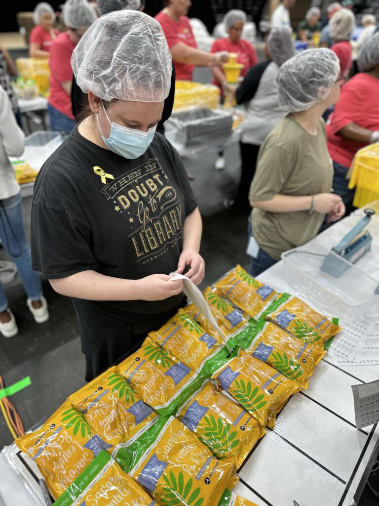 A person wearing a black t-shirt with the text "WHEN IN DOUBT GO TO THE LIBRARY" and a yellow ribbon pin is packing yellow and green bags labeled "Savory Lentils & Rice" on a table. The person is also wearing a hairnet and a face mask. Other people in the background are similarly dressed with hairnets and are engaged in packing activities.