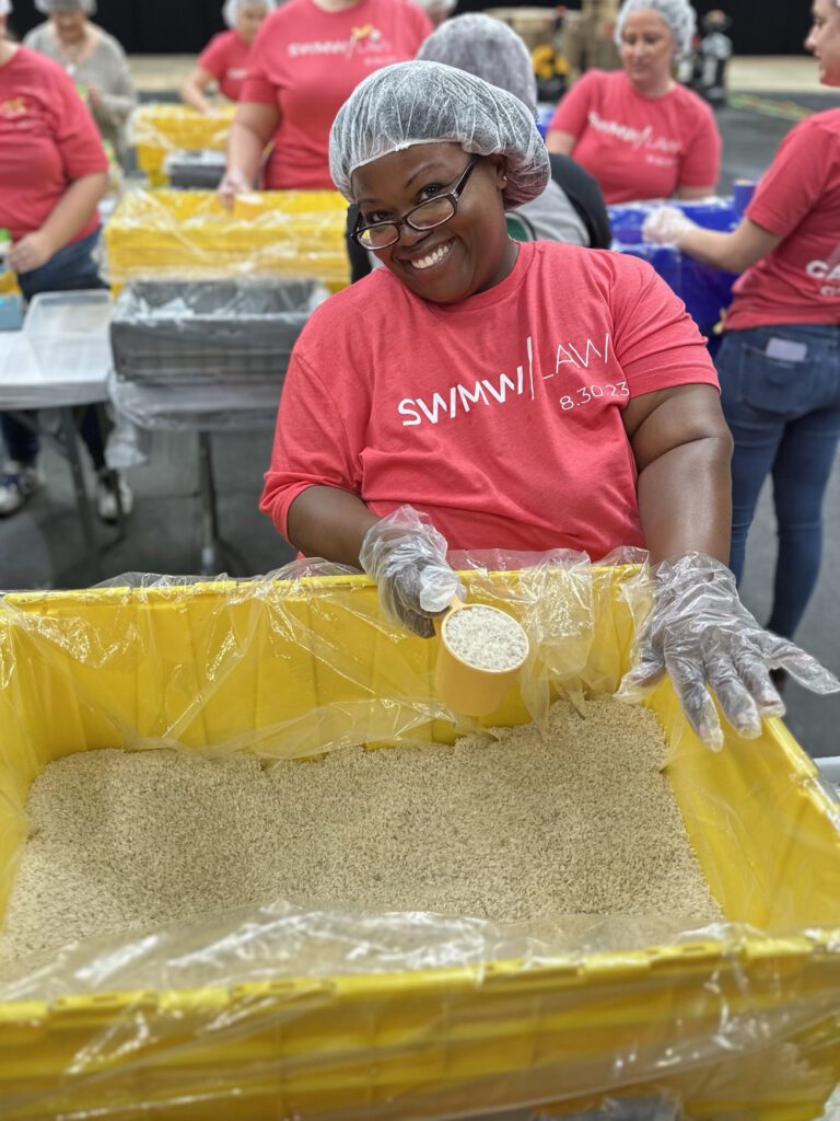 A smiling woman wearing glasses, a hairnet, and a red "SWIMW|LAW 8.30.23" t-shirt is scooping rice from a large yellow bin lined with plastic. She is wearing clear disposable gloves, and several other people in similar red shirts and hairnets are visible in the background, engaged in similar activities.