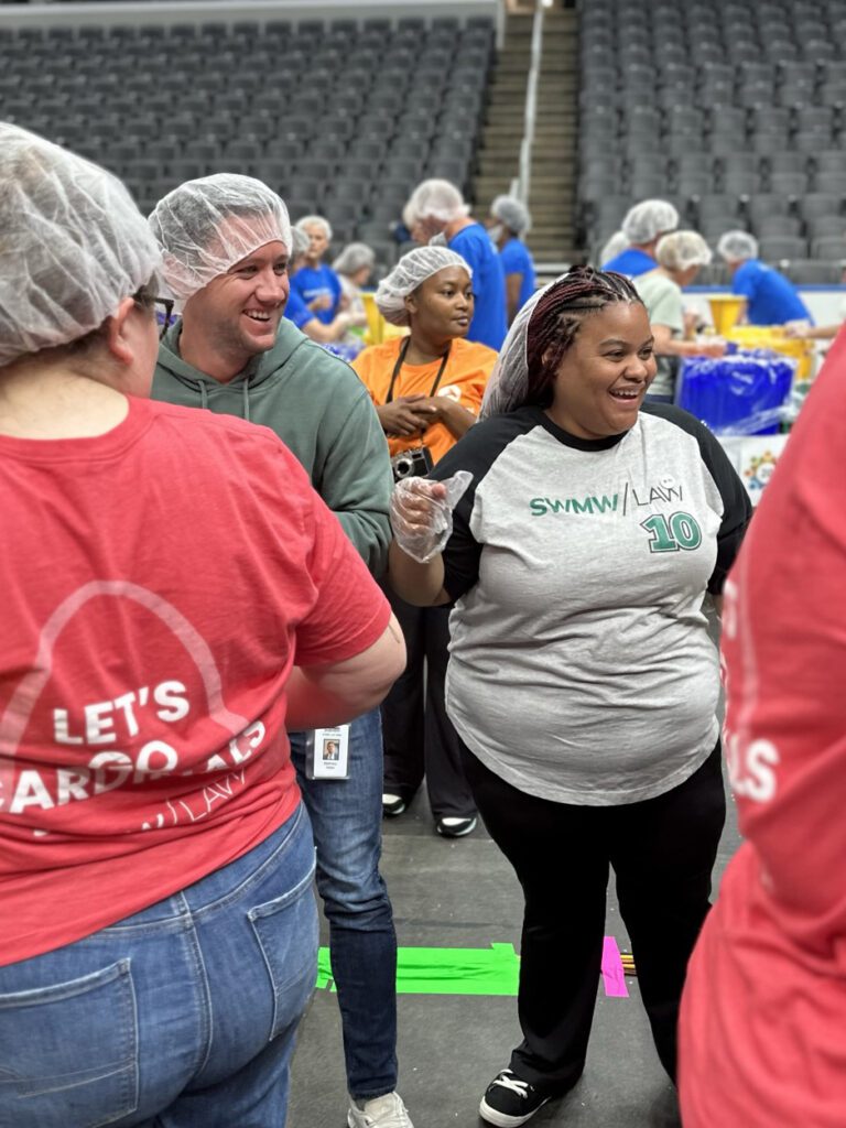 A group of people wearing hairnets and gloves are gathered indoors, likely participating in a volunteer or community event. One woman in the foreground is smiling and wearing a gray and black shirt with "SWMW / LANI 10" printed on it. Another person in a green hoodie and jeans is smiling and interacting with others. Several people in the background are wearing blue shirts and also hairnets, working at tables with yellow and blue containers. The setting appears to be a large venue with rows of empty gray seats.