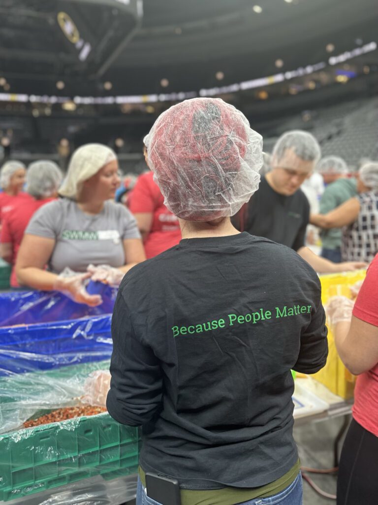 A group of people wearing hairnets and gloves are gathered around tables with large bins, likely participating in a food packaging or sorting event. The person in the foreground is wearing a black shirt with green text on the back that reads "Because People Matter." The setting appears to be indoors, possibly in a large hall or arena.