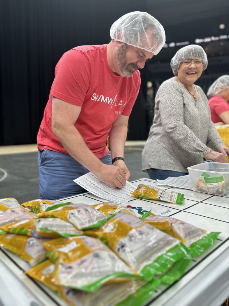 A man wearing a red "SWIMMY LAV" t-shirt and a hairnet is checking off items on a sheet of paper at a table filled with yellow and green packaged goods. Next to him, a smiling woman in glasses, a gray patterned blouse, and a hairnet is also working at the table, which has a plastic container with more packages and papers. The background is dark and out of focus.
