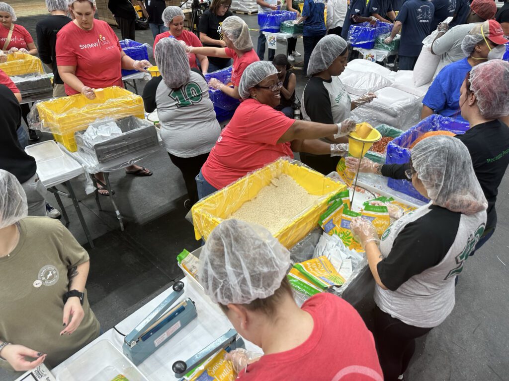A group of volunteers wearing hairnets and gloves are working together in an indoor setting to package food. They are scooping grains and lentils from large yellow bins into small plastic bags using funnels. The volunteers are standing around tables with various containers of food items and packaging materials. Some volunteers are wearing red shirts, while others wear gray and black shirts. The atmosphere appears organized and collaborative.
