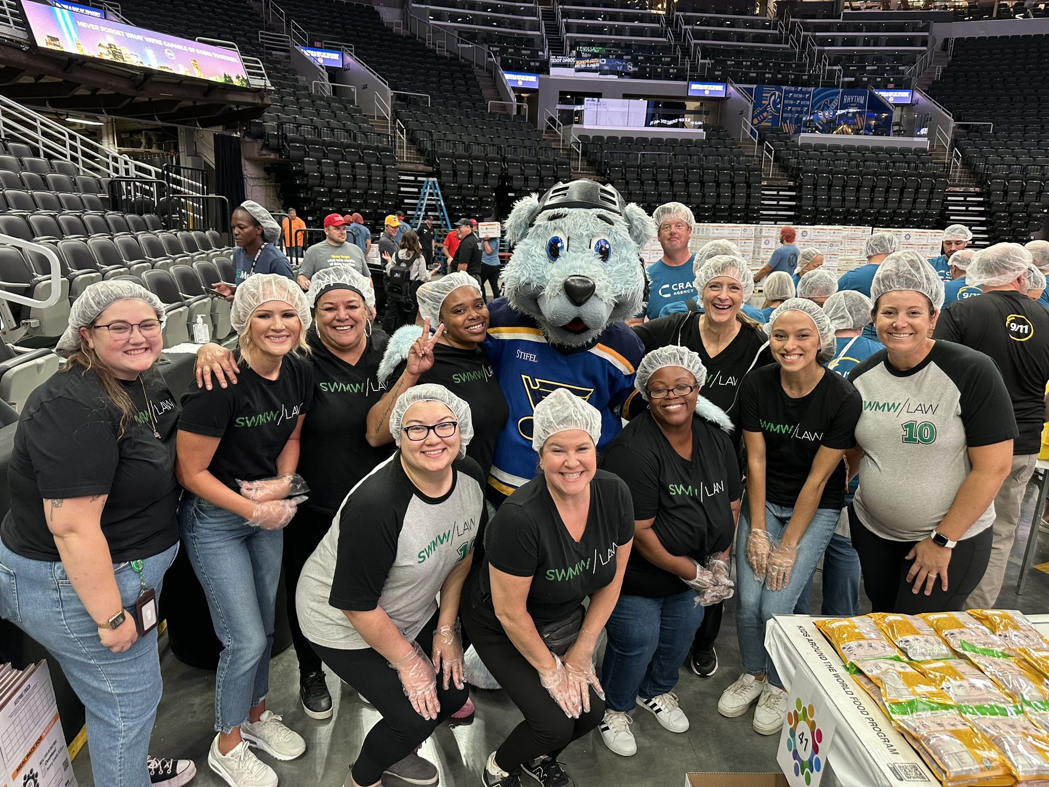 A group of people wearing hairnets and black or gray "SWMW | LAW" t-shirts pose together indoors on a sports arena floor. They are smiling and some wear plastic gloves. In the center is a person in a blue and yellow St. Louis Blues mascot costume. A table with packaged food items is visible in the foreground on the right. Empty stadium seats and other people are in the background.
