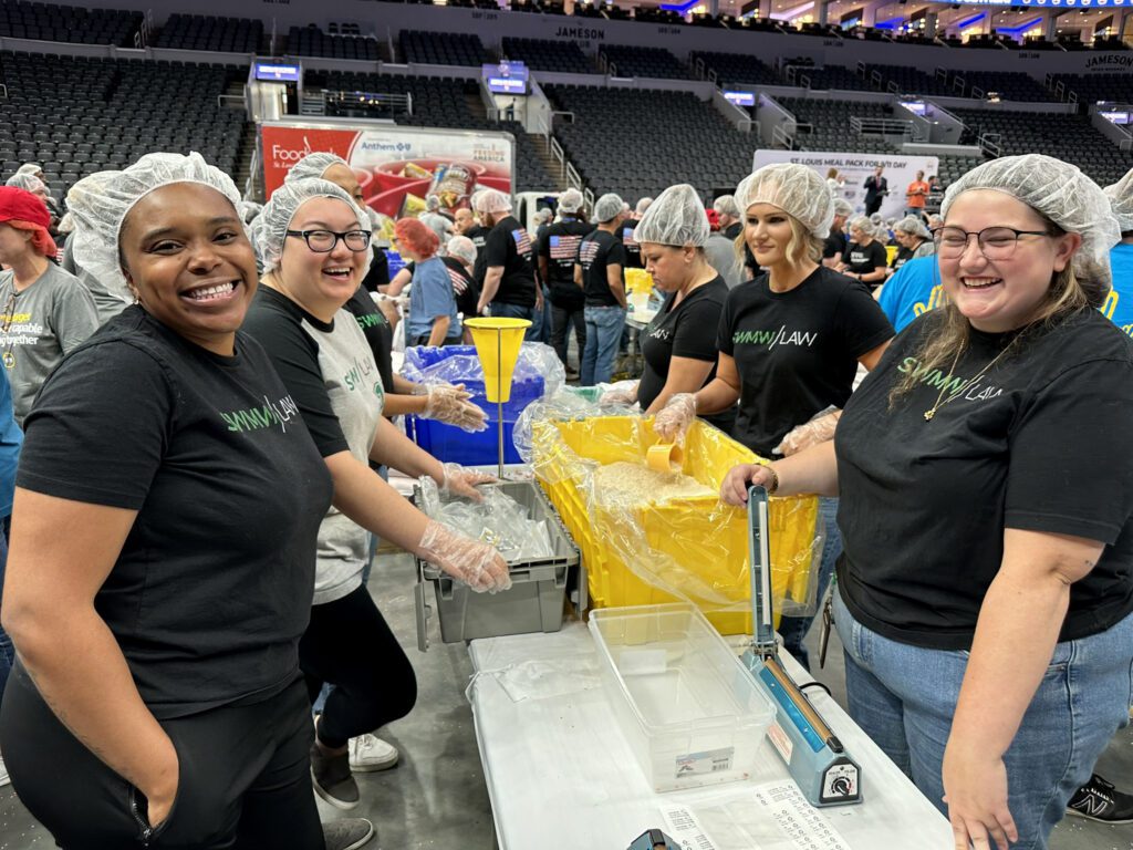 A group of people wearing hairnets and black or gray shirts with "SWMW LAW" printed on them are volunteering at a food packing event in an indoor arena. They are smiling and working together around tables with large yellow bins filled with rice and plastic containers, using scoops and sealing machines. The background shows more volunteers and empty stadium seats.