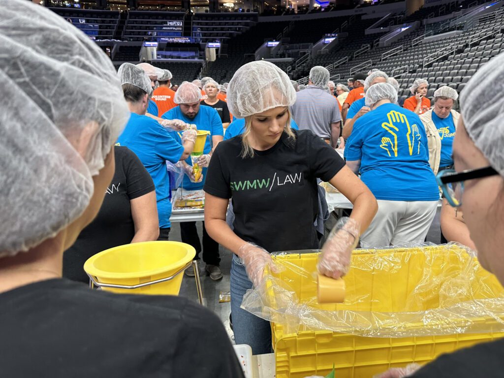 A group of people wearing hairnets and gloves are participating in a food packaging event inside a large indoor arena. The central woman, wearing a black "SWMW LAW" t-shirt, is scooping food into a large yellow container lined with plastic. Other volunteers in blue and orange shirts are working at tables in the background, filling smaller yellow buckets. The arena seats are mostly empty.