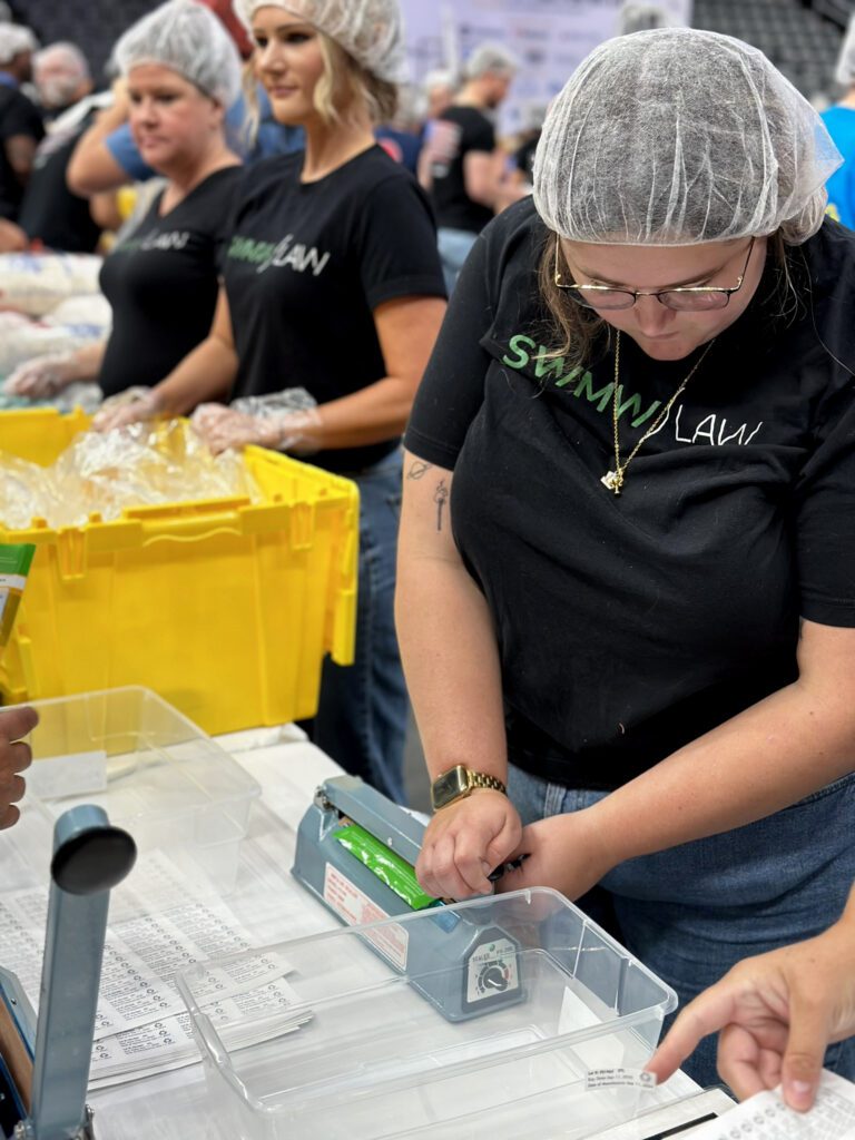 A group of people wearing black "Swann Law" t-shirts, hairnets, and plastic gloves are working at a table. One person in the foreground is sealing a green package with a heat sealer, while others in the background handle items near large yellow bins. The table has clear plastic containers and sheets with printed labels.