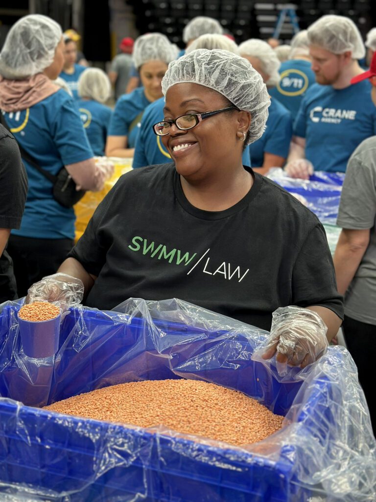 A smiling woman wearing glasses, a black "SWMW/LAW" t-shirt, a hairnet, and plastic gloves is scooping orange lentils from a large blue plastic bin lined with a clear plastic bag. Several other people in hairnets and blue shirts are visible in the background, participating in the same activity.
