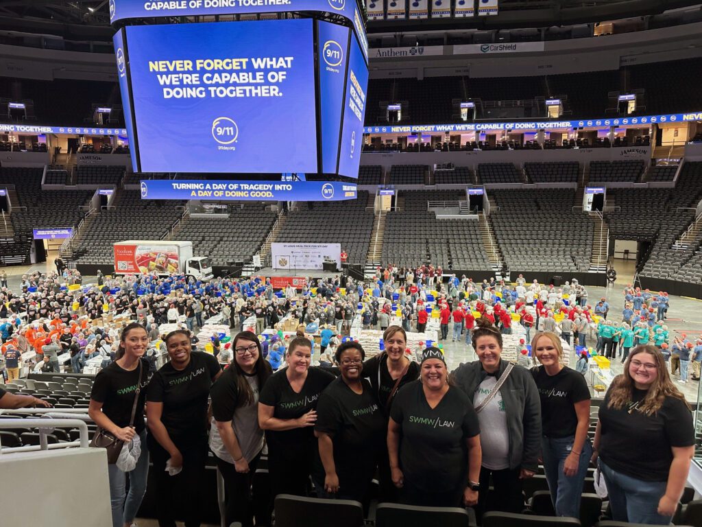 A group of ten people wearing black "SWMW LAW" t-shirts stand in the foreground of a large indoor arena. Behind them, hundreds of volunteers in colorful shirts are gathered around tables and boxes, participating in a large organized event. Above, a large digital screen displays the message "NEVER FORGET WHAT WE'RE CAPABLE OF DOING TOGETHER" with a 9/11 logo, and a scrolling banner reads "TURNING A DAY OF TRAGEDY INTO A DAY OF DOING GOOD." The arena seating is mostly empty.