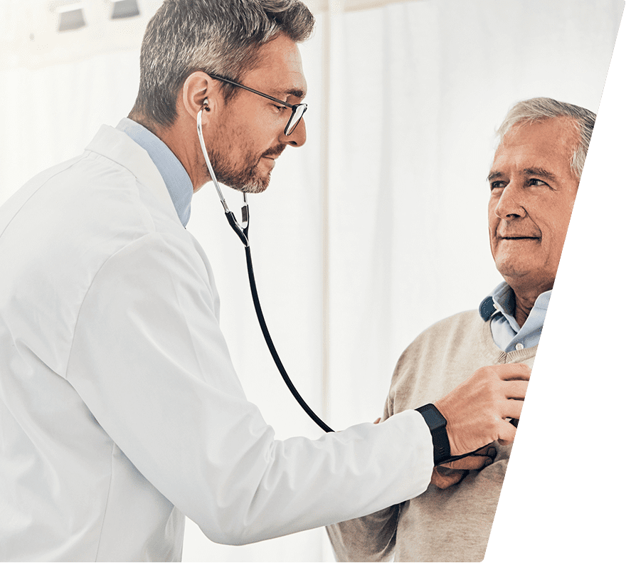 A male doctor wearing glasses and a white coat is using a stethoscope to listen to the chest of an older male patient who is wearing a beige sweater over a blue collared shirt. The doctor is focused, and the patient looks calm. The background is light and neutral.